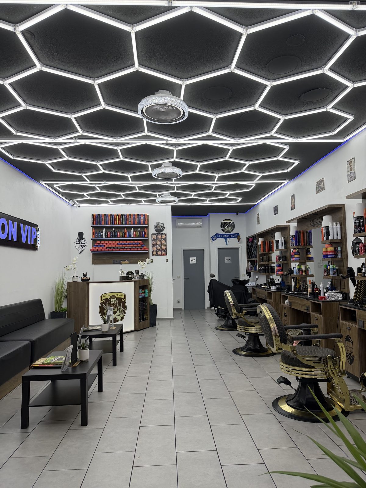 Blick der Länge nach durch den Salon VIP: Wabenlicht an der Decke, goldene Barberstühle rechts, Wartesofa links